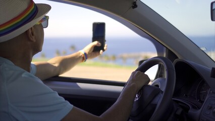 Young adult gay tourist with bracelet, sunglasses and rainbow hat photographs coastal landscape from rental car. LGBT tourism concept