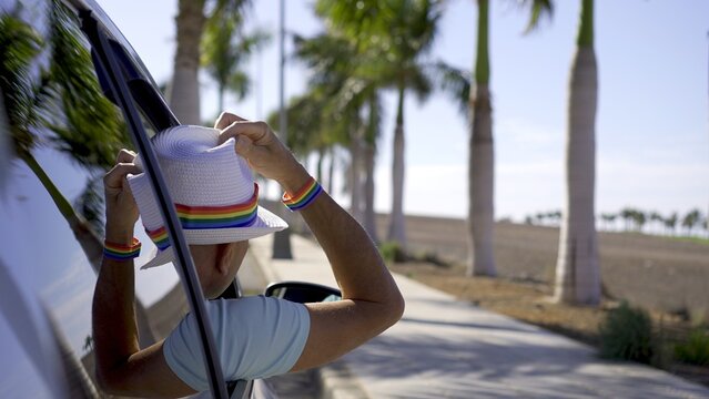 Gay traveler wearing hat and rainbow bracelet leans out of car window to look at tropical paradise. Gay tourism concept