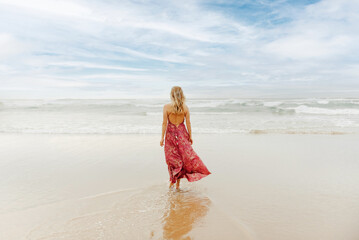 Young blonde woman with her back turned dressed in summer dress walking alone along the shore of the beach towards the sea.