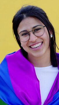 Young lesbian woman smiling at camera over yellow background celebrating LGBTQ pride day festival covered with rainbow flag proud to be homosexual. Vertical HD videO