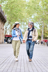 Vertical. Happy adult Caucasian tourist couple looking at other walking holding hand for city street. Mature love people enjoying romantic getaway. Positive relationships and spring retiree holidays