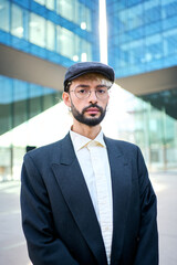 Vertical video. Young Caucasian hipster business man in a smart blue suit, hat and glasses standing serious and confident in front of an office building, adjusts jacket ready for a formal work event