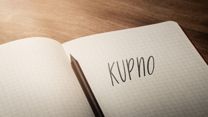 A handwritten inscription "Kupno" on a grille of an open notebook on a wooden countertop, next to a black pencil, lighting of light. (selective focus), translation: purchase