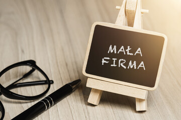 Black writing board on a wooden frame with the inscription "Mała Firma", next to black glasses and a pen (selective focus) translation: small company