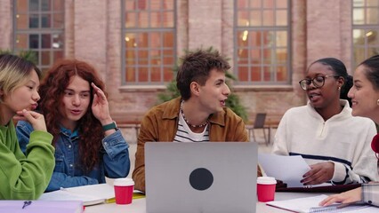 Group of smiling and concentrated diverse students using laptop and talking doing brainstorming on university campus. Young people friends sitting working and studying together outdoor