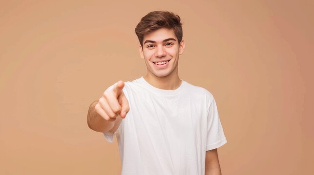 A young man wearing a plain white T-shirt against a light brown background is posing pointing at the camera.