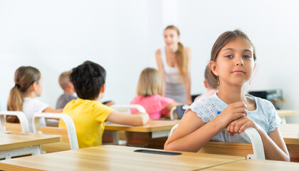 Portrait of positive tween schoolgirl sitting at school desk at lesson in class, looking confidently at camera