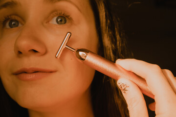 Obraz premium Young beautiful white woman, girl is using vibrating golden facial massager on black background. Face massage. Natural beauty youth Female skin care.