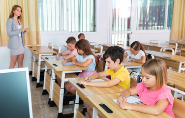 Fototapeta premium Upset serious preteen boy and girl listening to teacher and writing exercises in notebook at lesson in primary school