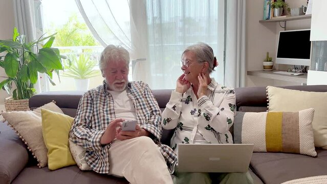 Senior couple sitting on sofa at home using laptop and cell phone discussing diverging on a topic, 70 year old Caucasian seniors sitting in living room browsing, shopping online, following social medi