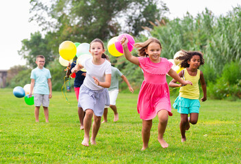 Multiracial group of cheerful tweenagers holding colored balloons in hands, running together in summer city park. Happy childhood concept