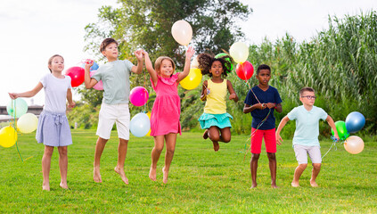 Portrait of cheerful tween girls and boys holding colorful balloons in hands jumping together in...