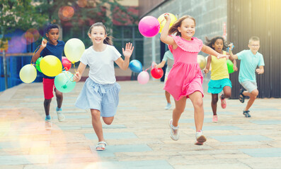Young joyful boys and girls running through city streets with balloons in hands.