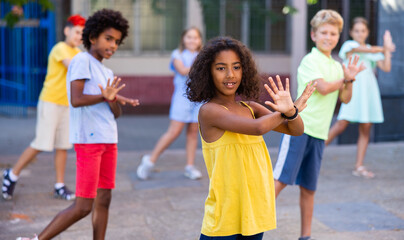 Smiling curly african american preteen girl dancing modern choreography with group of tweenagers on city street on summer day..