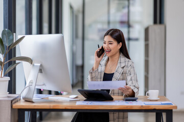 Asian woman entrepreneur busy with her work in the office. Young Asian woman talking over phone or cellphone while sitting at her desk.
