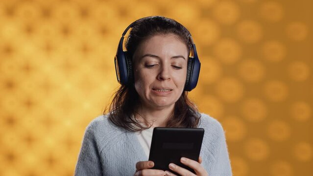 Woman wearing headphones, practicing new language vocabulary using online app on tablet, isolated over studio background. Lady speaking foreign phrases, reading them on device screen