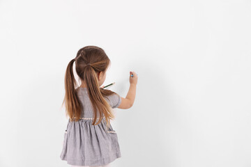 Little girl drawing on white wall indoors, back view. Space for text