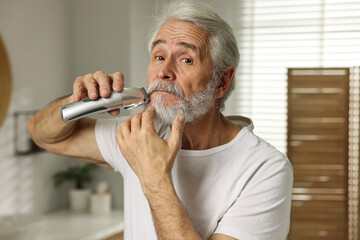 Senior man trimming beard with electric trimmer in bathroom - Powered by Adobe