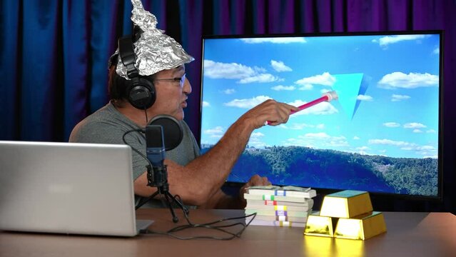 A crazy UFO conspiracy theorist with a tin foil hat records a podcast in his home studio. UFO video on monitor. UFO footage from my portfolio.  	