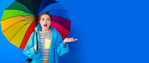Surprised young woman in raincoat and with umbrella showing something on blue background with space for text