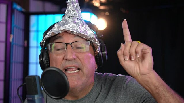 A crazy UFO conspiracy theorist with a tin foil hat records a podcast in his home studio. Talks directly to camera.  	