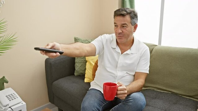 Mature man in casual attire leisurely holding a mug and a remote control in a cozy living room setting.