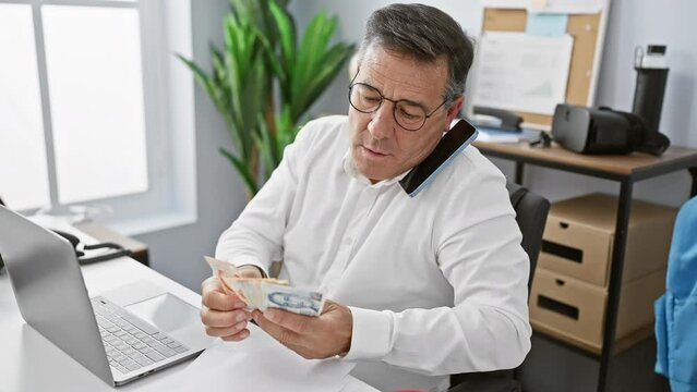Mature businessman counts singaporean dollars multitasking with phone at desk in office