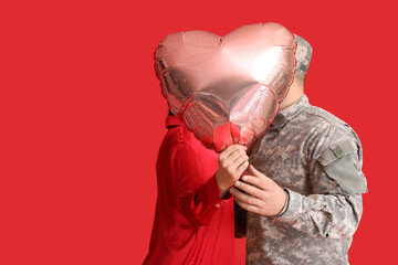 Man in military uniform and his wife kissing behind heart-shaped balloon on red background....