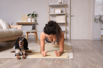 Sporty young African-American woman with cocker spaniel doing plank at home