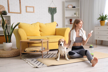 Sporty young woman with cute Beagle dog and mobile phone after doing yoga on mat at home