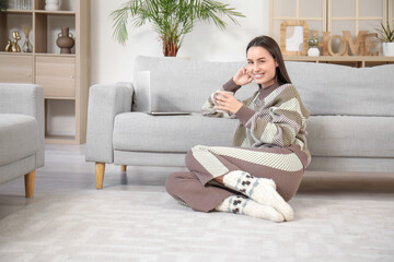 Obraz premium Pretty young woman with cup of tea and laptop on grey sofa in living room