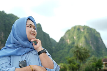 Asian model in hijab wearing blue shirt is posing in nature with mountains and water in the countryside as a backdrop