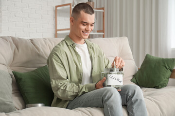 Male student putting money in jar bank on sofa in room. Concept of savings for education
