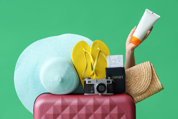 Female hand with sunscreen cream, beach accessories, passport and suitcase on green background,...