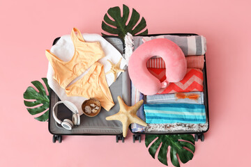 Suitcase with beach accessories, headphones, palm leaves and decor on pink background
