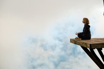 A young girl of European appearance meditates and does yoga on a wooden platform in the mountains,...