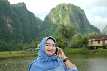 Asian model in hijab wearing blue shirt is posing in nature with mountains and water in the countryside as a backdrop