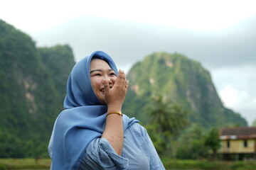 Asian model in hijab wearing blue shirt is posing in nature with mountains and water in the...