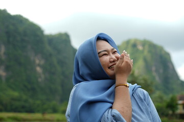 Asian model in hijab wearing blue shirt is posing in nature with mountains and water in the countryside as a backdrop