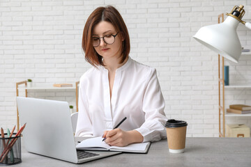 Young businesswoman writing on notebook and sitting at table with modern laptop in stylish office