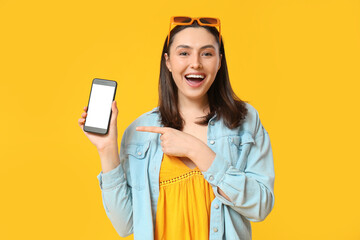 Beautiful young happy woman in stylish denim jacket pointing at mobile phone on yellow background