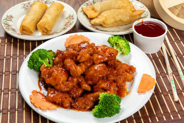 A plate of Chinese General Tso's chicken with plates of egg rolls and spring rolls in the background