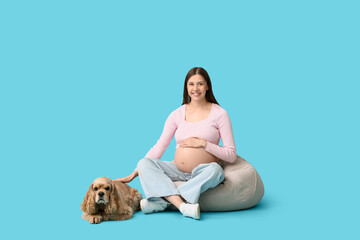 Obraz na płótnie Canvas Beautiful young pregnant woman with cute Cocker Spaniel dog sitting on beanbag chair against blue background