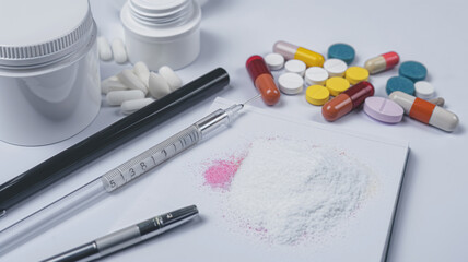 Medical syringe, pills, capsules, and spilled powder on white surface.