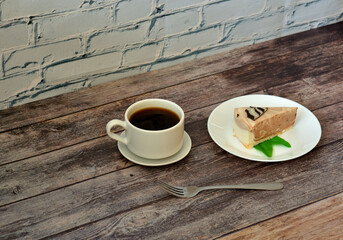 A plate with a slice of cream cheesecake with mint and a cup of hot black coffee on a light wooden...