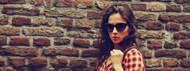 Beautiful brunette woman with long brown hair in fashion black sunglasses standing on the old red brick wall building background in casual red shirt in spring city. Closeup vintage banner portrait