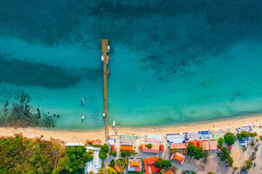 Aerial top view of colorful umbrellas on sandy beach, swim in blue sea summer sunny day. Tropical island sandy azure bay pier village buildings settlement surrounded exotic green forests.