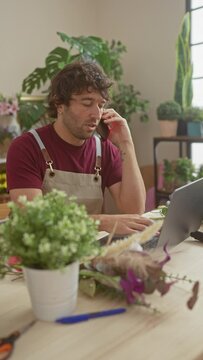 Hispanic man with a beard talking on the phone and using a laptop in a flower shop interior.