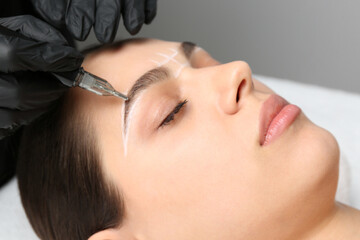 Beautician making permanent eyebrow makeup to young woman on grey background, closeup