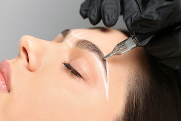 Beautician making permanent eyebrow makeup to young woman on grey background, closeup